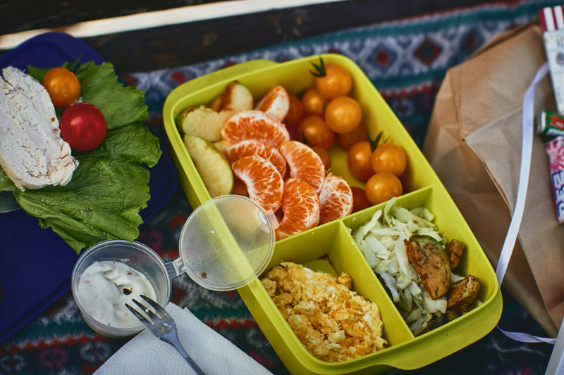 Colorful bento lunchbox with organized compartments containing fresh fruits, vegetables, and rice arranged on a decorative patterned tablecloth