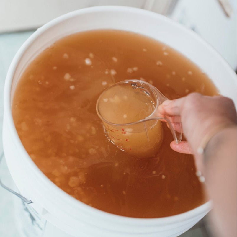 Hand holding measuring cup pouring brown liquid into large white container for beverage production