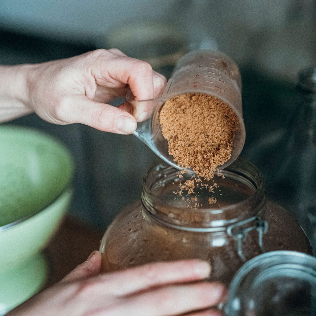 Person pouring brown powder from a transparent container into a glass jar for beverage or food preparation