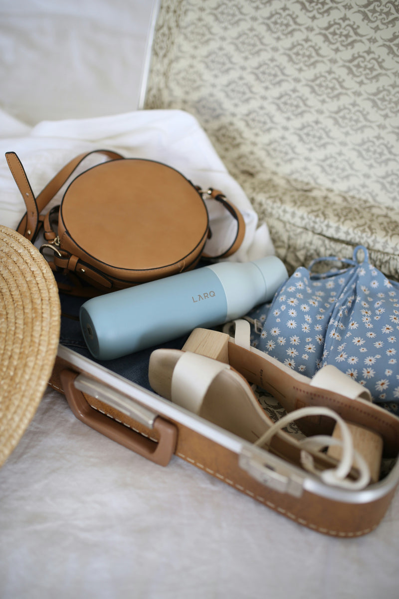 Compact travel suitcase with blue water bottle, brown bag, and floral fabric arranged on textured surface - travel essentials flatlay