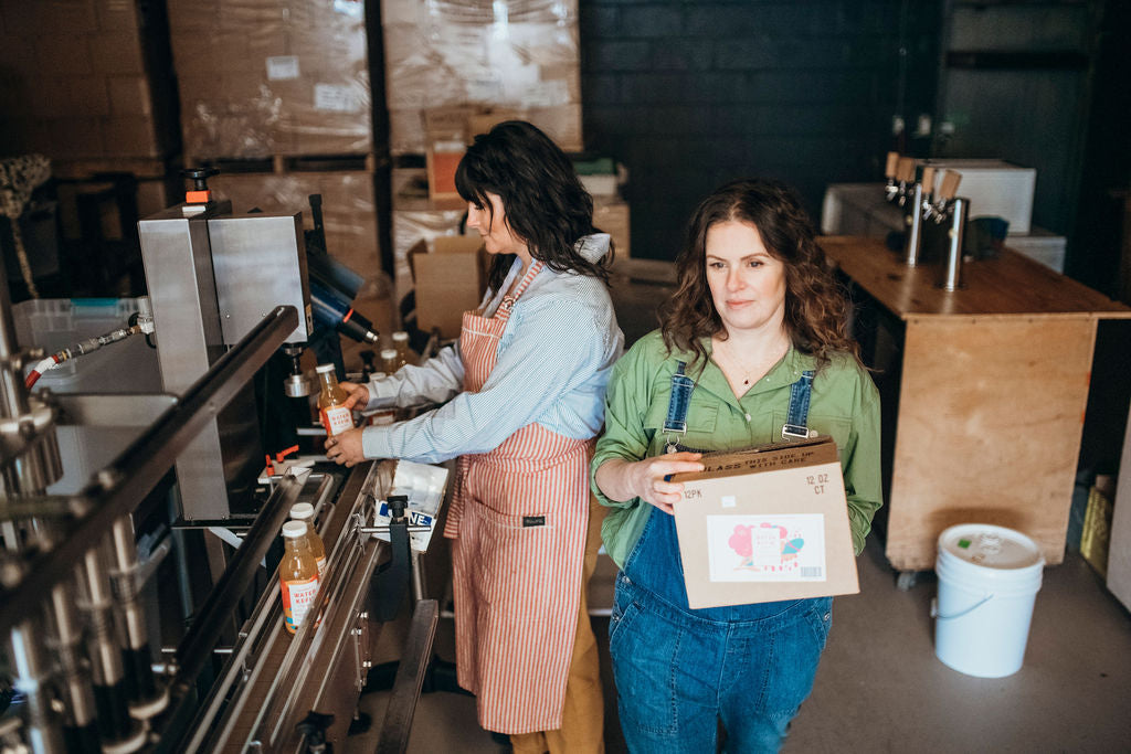 Two women in kitchen or workshop setting, one woman holding colorful product packaging box