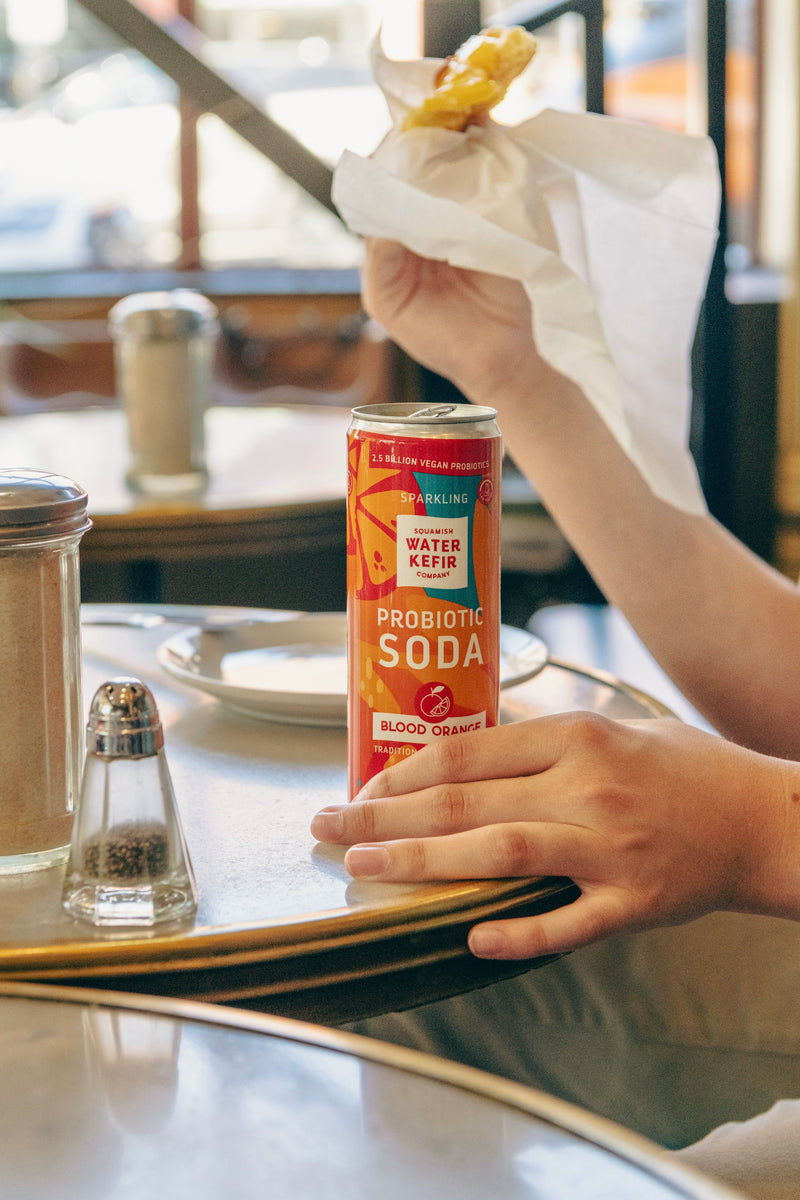 Person holding a can of probiotic soda at a table with a blurred background